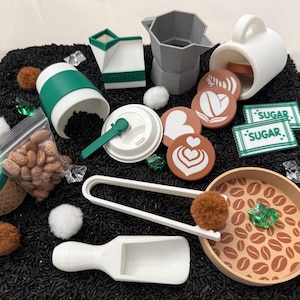 May include: A collection of coffee-themed toys, including a white mug, a gray moka pot, and a green and white coffee cup. There are also coffee beans, sugar packets, and latte art coasters. The items are arranged on a black surface.