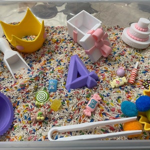 May include: A sensory bin filled with white rice and colorful sprinkles. The bin includes a purple crown, a purple number four, a white gift box with a pink bow, a white cake with pink frosting and the words "Happy Birthday", a purple bowl, a white scoop, a pair of white tongs, a yellow candy, a pink candy, a white candy, a yellow gummy, a pink candy with a cherry on top, a white candy with a bunny on it, a pink and white candy, a white and yellow candy, a red and white candy, a brown candy, and a yellow container with colorful pom-poms.