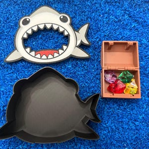 A Shark container with the lid off, and a small treasure chest filled with colorful gems. The items are arranged on blue rice.