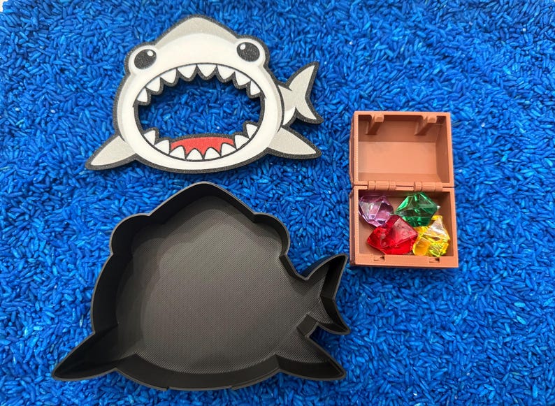 A Shark container with the lid off, and a small treasure chest filled with colorful gems. The items are arranged on blue rice.