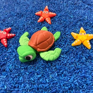 A playful scene featuring a green turtle with a brown shell, surrounded by three colorful starfish. The starfish are red and yellow, with white accents. The toys are set on a bed of blue rice.