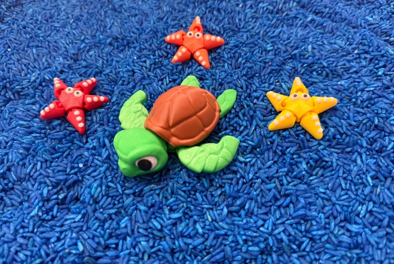 A playful scene featuring a green turtle with a brown shell, surrounded by three colorful starfish. The starfish are red and yellow, with white accents. The toys are set on a bed of blue rice.