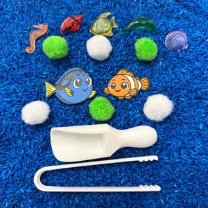 An ocean-themed sensory bin with blue rice, featuring toy fish, a seahorse, a seashell, green pom-poms, and white scoops and tongs. The toys are arranged on blue rice, creating a playful and engaging scene.