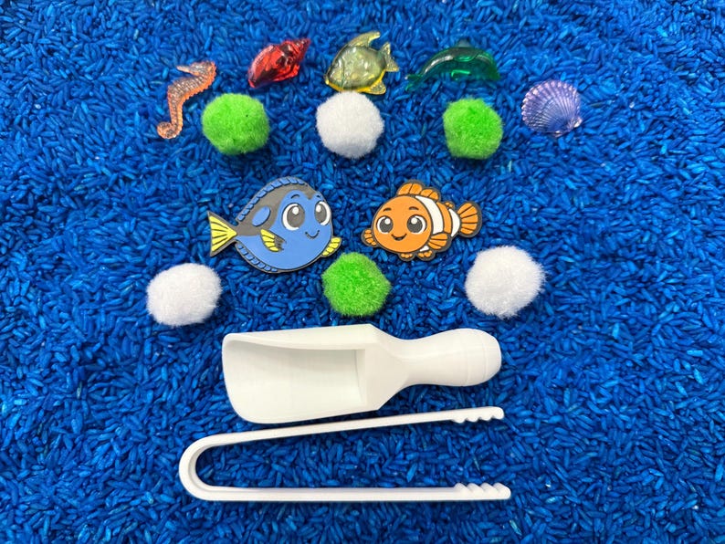 An ocean-themed sensory bin with blue rice, featuring toy fish, a seahorse, a seashell, green pom-poms, and white scoops and tongs. The toys are arranged on blue rice, creating a playful and engaging scene.