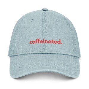 Puede incluir: Gorra de béisbol de mezclilla azul claro con la palabra "caffeinated." bordada en rojo. La gorra tiene una visera curva y un botón en la parte superior. Un accesorio casual.