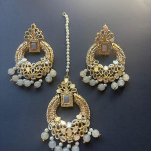 May include: A gold-toned jewelry set with a large, round, mirrored pendant and matching earrings. The pendant and earrings are adorned with small, mirrored pieces and white beads.