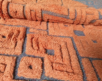 May include: Close-up of a textured, orange rug with a geometric pattern. The rug has a plush, looped pile and a teal background. The design features repeating square and rectangular shapes.