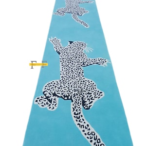 May include: A light blue runner rug featuring a repeating pattern of stylized leopards in black and white. The leopards are depicted in a playful, leaping pose, creating a dynamic and eye-catching design. The rug is a long, rectangular shape.