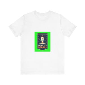 Peut inclure: Un t-shirt blanc à manches courtes avec un motif rectangulaire centré sur un fond vert vif. Le motif représente une fusée de dessin animé au décollage avec le texte "DEPLOY DREAMS" et des symboles de code comme des crochets et des barres.