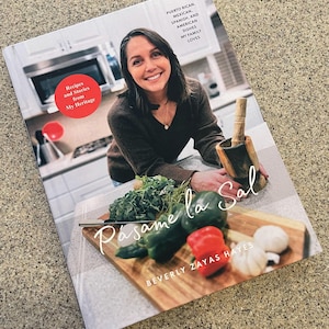 Könnte beinhalten: Ein Kochbuch mit dem Titel "Pasame la Sal" von Beverly Zayas Hayes. Das Cover zeigt eine Frau in einem braunen Pullover, die einen Mörser und einen Stößel hält, im Vordergrund eine hölzerne Schneidebrett mit frischem Gemüse und Knoblauch. Der Text auf dem Cover lautet "Rezepte und Geschichten aus meinem Erbe" und "Puertoricanische, mexikanische, spanische und amerikanische Gerichte, die meine Familie liebt."