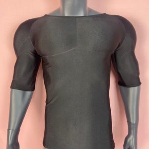 Puede incluir: Camiseta de compresión negra con hombros y brazos acolchados, exhibida en un maniquí. La camiseta tiene cuello redondo y mangas tres cuartos. La tela parece suave y ajustada, diseñada para una apariencia esculpida.