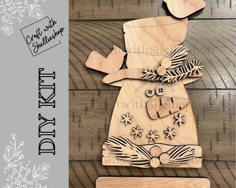 May include: A wooden DIY kit for a snowman decoration. The kit includes laser-cut wooden pieces for the snowman's body, hat, and decorative elements like birds, buttons, and snowflakes. The text "DIY KIT" is visible.