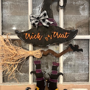 May include: A hanging Halloween decoration featuring a black witch hat with a purple band, a black and white buffalo check bow, and the orange text trick or treat. Below, a broom with straw bristles and two black boots with purple accents.