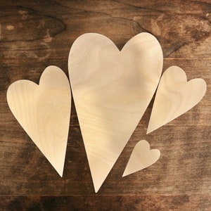 May include: Four unfinished wooden heart cutouts of varying sizes are arranged on a dark wood surface. The hearts are light-colored, with visible wood grain. The largest heart is approximately 10 inches tall, while the smallest is about 2 inches.