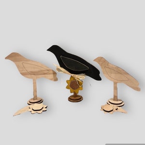 May include: Three wooden bird cutouts, two are light brown and one is black. The birds are standing on a wooden base with a sunflower design.