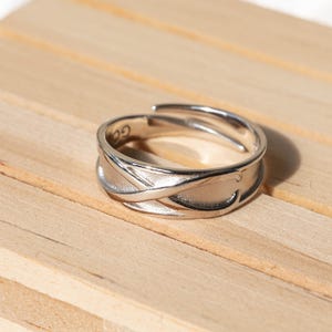 Puede incluir: Un anillo plateado con un diseño intrincado e entrelazado. El anillo tiene una superficie texturizada y una pequeña inscripción. El anillo se muestra sobre una superficie de madera clara.