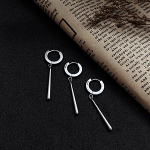 May include: Three silver hoop earrings with a long, thin, silver dangle. The earrings are on a black background.