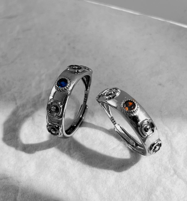 May include: Two silver rings with floral designs and small gemstones. One ring has a blue gemstone and the other has an orange gemstone.