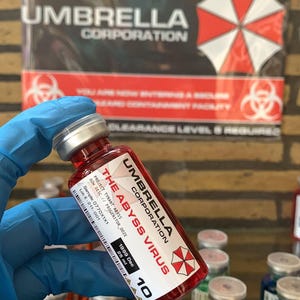 Może przedstawiać: Mała szklana fiolka wypełniona czerwonym płynem, z białą etykietą z napisem UMBRELLA CORPORATION, THE ABYSS VIRUS, 25 ml oraz czerwono-białym logo parasola. Fiolka posiada srebrną metalową zakrętkę oraz nadrukowany kod kreskowy na etykiecie.