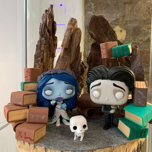 May include: A hand-crafted diorama featuring a bride and groom from the movie 'The Corpse Bride'. The figures are surrounded by stacks of books, a small skeletal dog, and a wooden base.