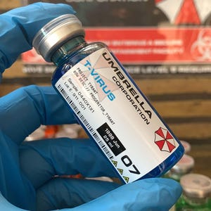 Op de afbeelding: Een flesje met het label "T-VIRUS" van Umbrella Corporation, met blauwe vloeistof. Het label bevat informatie over Project Tyrant, een sample code en 25 ml volume. Het flesje wordt vastgehouden door iemand met blauwe handschoenen.