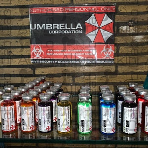 Può includere: Un'esposizione di fiale colorate con l'etichetta "Umbrella Corporation" e il logo Umbrella. Le fiale sono disposte su una mensola di vetro, con un cartello sopra che recita "Solo personale autorizzato". Il cartello presenta anche il logo Umbrella e simboli di pericolo biologico.