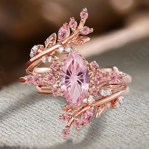 May include: A rose gold ring featuring a central marquise-cut pink gemstone surrounded by smaller pink and clear stones. The band is designed with delicate leaf-like metalwork extending from the center, accented with additional pink and clear stones.