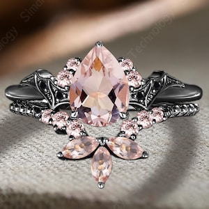 Może przedstawiać: Czarny pierścionek zaręczynowy z gruszkowym, brzoskwiniowym kamieniem szlachetnym jako centralnym elementem. Pierścionek jest ozdobiony mniejszymi różowymi kamieniami szlachetnymi i dekoracyjnym filigranem. Pierścionek leży na neutralnej powierzchni.