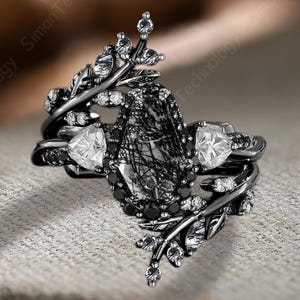 May include: A black ring set with a central, rectangular, black and white stone, flanked by two triangular white stones. The ring features a leaf-like design with small white accents, set against a neutral background. The ring is a piece of jewelry.