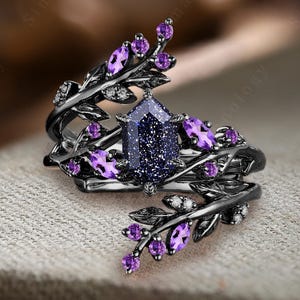 May include: Black ring with a large blue gemstone surrounded by smaller purple gemstones and black leaves. The ring is on a purple fabric background.