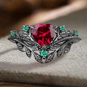 May include: Black ring featuring a large heart-shaped red gemstone, surrounded by smaller green gemstones and intricate leaf designs.