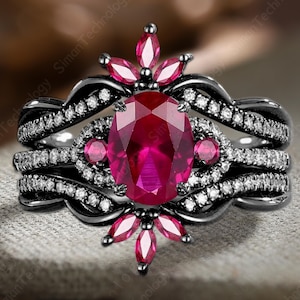 Może przedstawiać: Trzyczęściowy zestaw pierścionków z dużym owalnym rubinem w środku, otoczonym mniejszymi rubinowymi akcentami i białymi kamieniami szlachetnymi. Czarny metalowy pierścień ma skręconą konstrukcję. Pierścionek jest osadzony na neutralnym tle z tkaniny.