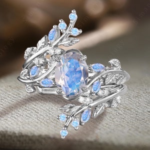Puede incluir: Un anillo de plata con una gran piedra preciosa ovalada azul rodeada de pequeñas piedras preciosas transparentes. El anillo presenta un diseño delicado de ramas entrelazadas con hojas y pequeñas piedras preciosas azules.