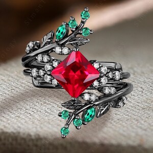 Puede incluir: Un anillo negro con una gran piedra roja cuadrada rodeada de piedras blancas más pequeñas. El anillo tiene un diseño de hojas con piedras verdes en la banda.
