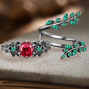 May include: A two-piece ring set featuring a central red gemstone ring and a separate, spiraling band with green leaf-shaped accents. The rings have a dark, possibly oxidized, metal finish, and the gemstones are set in floral designs.
