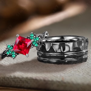 May include: A black ring set with a red square gemstone and green accents. The ring is on a brown surface.