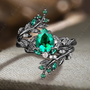 May include: A black ring set with a pear-shaped emerald green gemstone surrounded by smaller clear gemstones. The ring features a delicate black twig design with emerald green leaves.