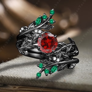 Puede incluir: Anillo negro con una gran piedra roja rodeada de piedras blancas más pequeñas. El anillo presenta un diseño de metal negro con dos ramas frondosas con piedras verdes.