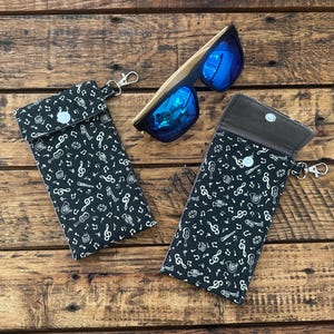 May include: Two black fabric eyeglass cases with a white musical note pattern, snap closures, and silver keychains. Sunglasses with blue lenses and wooden frames are on a wooden surface.