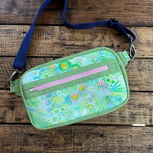 May include: A green fanny pack with a colorful geometric pattern, featuring a pink zipper and a navy blue strap. The bag has a rectangular shape with rounded corners and silver-colored metal hardware.