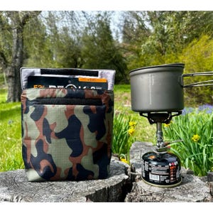May include: A camouflage-patterned pouch with a black zipper and a black strap. The pouch is open and a small orange and black label is visible inside. The pouch is sitting on a tree stump next to a small camping stove with a pot on top.