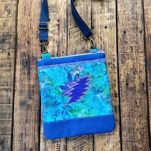 May include: A blue and turquoise crossbody bag with a blue lightning bolt design. The bag has a blue adjustable strap and a zipper closure. The bag is made of fabric with a floral pattern.