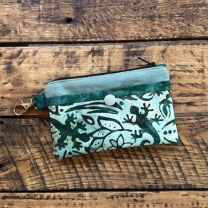 May include: A small, teal and green fabric coin purse with a lizard and leaf pattern. The purse has a black zipper, a silver-colored clasp, and a white button closure. The background is a wooden surface.