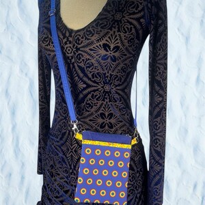 May include: A blue crossbody phone bag with a yellow and blue patterned design. The bag has a blue strap and a yellow trim. The bag is hanging on a dark blue patterned dress.