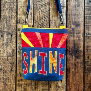 May include: A blue crossbody bag with a vibrant design. The top section features a red and yellow sunburst pattern, while the lower section displays the word "SHINE" in large, colorful letters. The bag has a yellow zipper and a blue strap.