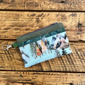 May include: A small, rectangular coin purse with a mountain and forest print. The purse has a light blue background with a zipper closure, a green accent, and a silver clasp. The design features trees and mountains.
