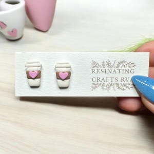 May include: A pair of white coffee cup earrings with a pink heart on each cup. The earrings are on a white card with the text "RESINATING CRAFTS RVA".