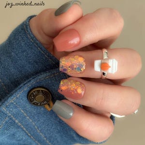 May include: Close-up of a hand with manicured nails. The nails feature a mix of colors and designs, including peach, glitter, and gray. A silver ring with a coffee cup design is also visible. The text "joz_winked_nails" is in the upper left corner.