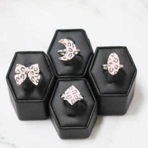 May include: Four rings with silver bands and pink leopard print designs are displayed on black stands. The ring designs include a bow, a crescent moon, a teardrop, and an oval shape. The rings are set against a white background.