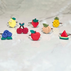 May include: A collection of fruit-themed rings, including a yellow lemon, red strawberry, and a slice of watermelon. Each ring features a different fruit design in vibrant colors, set on a silver-tone band. The rings are arranged on a white marble surface.